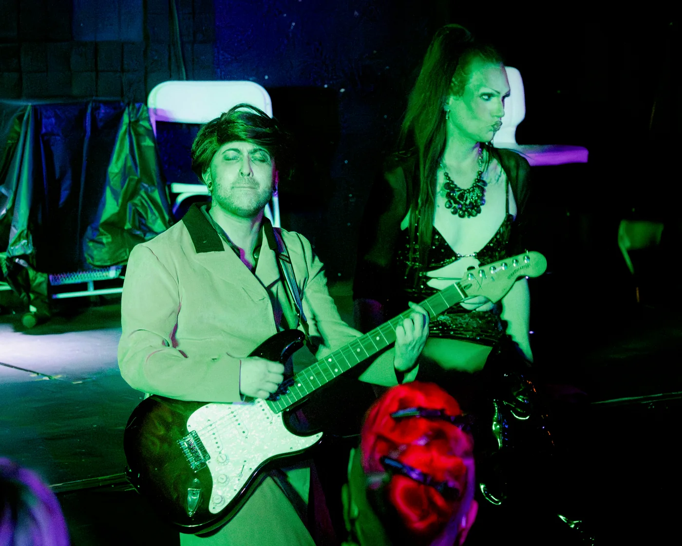 A Queen-y Todd performer plays an electric guitar beside a castmate under green stage lighting.